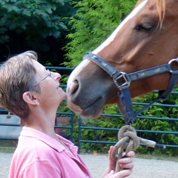 Joan holding a horse's bridle with her chin resting on the horse's muzzle and smiling up at it