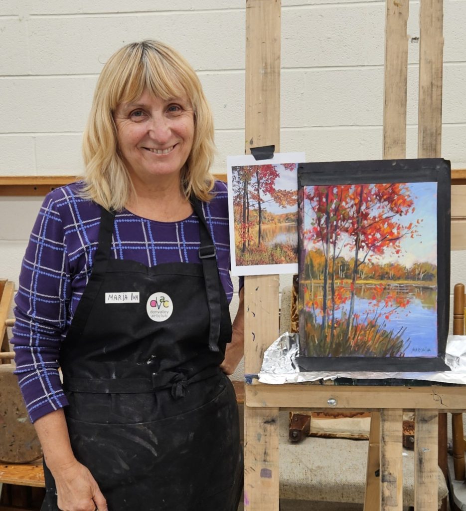 Maria Iva wearing an apron standing in front of her painting on an easel