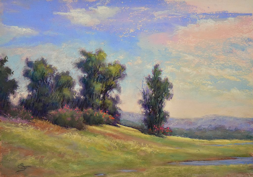 A grassy hill with trees next to a small pond with warm blues and pinks in the clouds in the sky